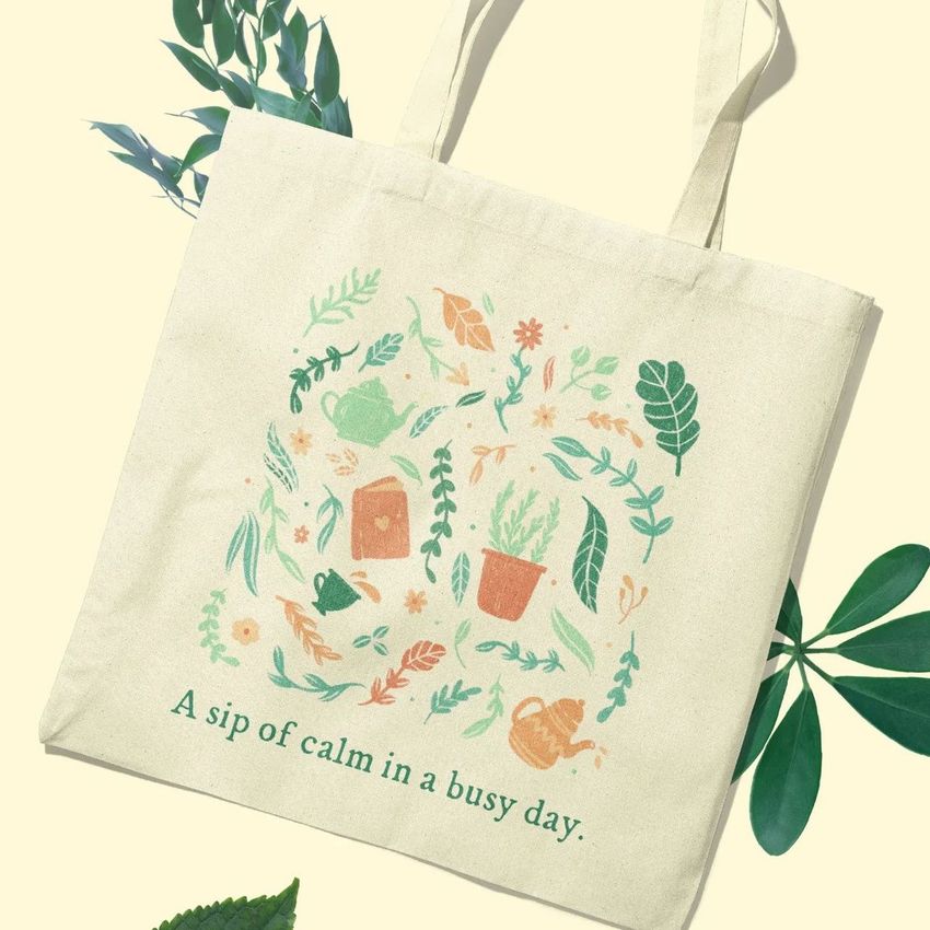 Tote bag mockup featuring a cream canvas bag with botanical illustrations and a teapot design, photographed on a pale background.