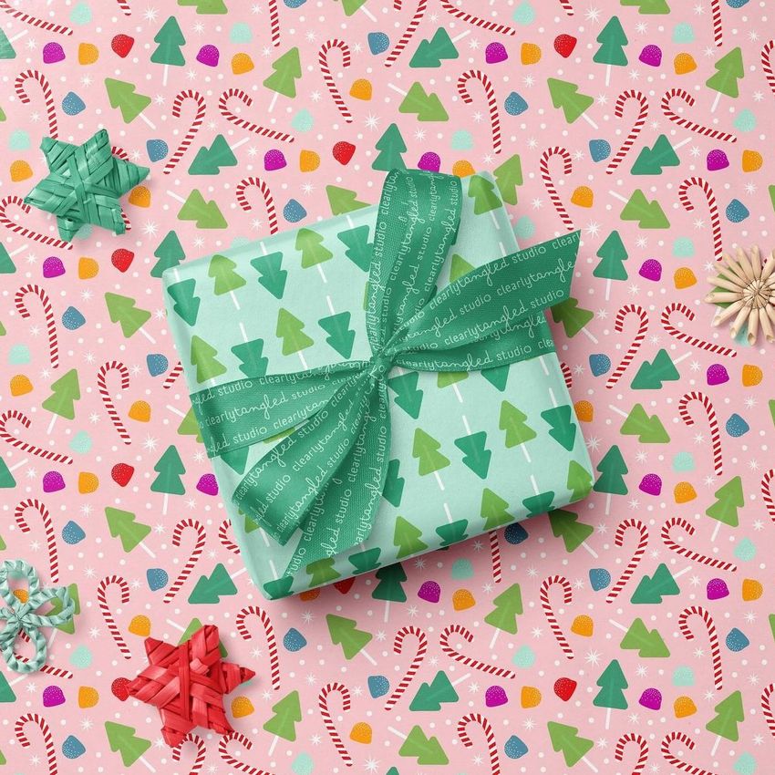 Square gift box mockup wrapped in green paper with tree pattern and ribbon, placed on colorful Christmas-themed wrapping paper.
