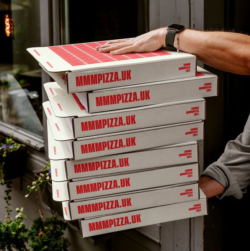 Stack of six white pizza boxes with red text, held outdoors by a person’s arm near a building entrance and potted plants.