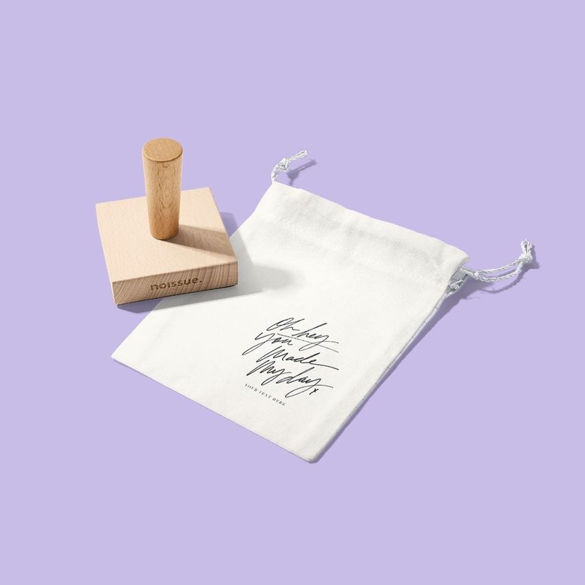 Wooden stamp mockup set on a light purple background, featuring a square wooden stamp and a white drawstring pouch with script text.