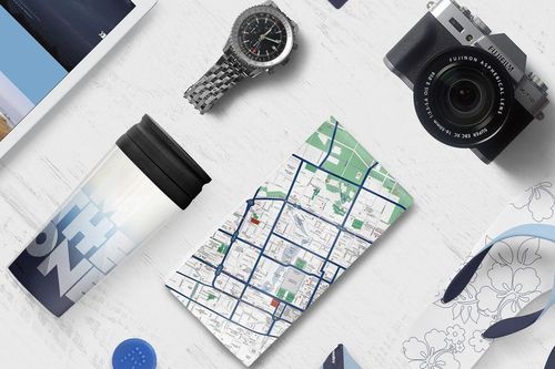Travel identity mockup with a folded city map, stainless steel watch, camera, and tumbler arranged on a light table.