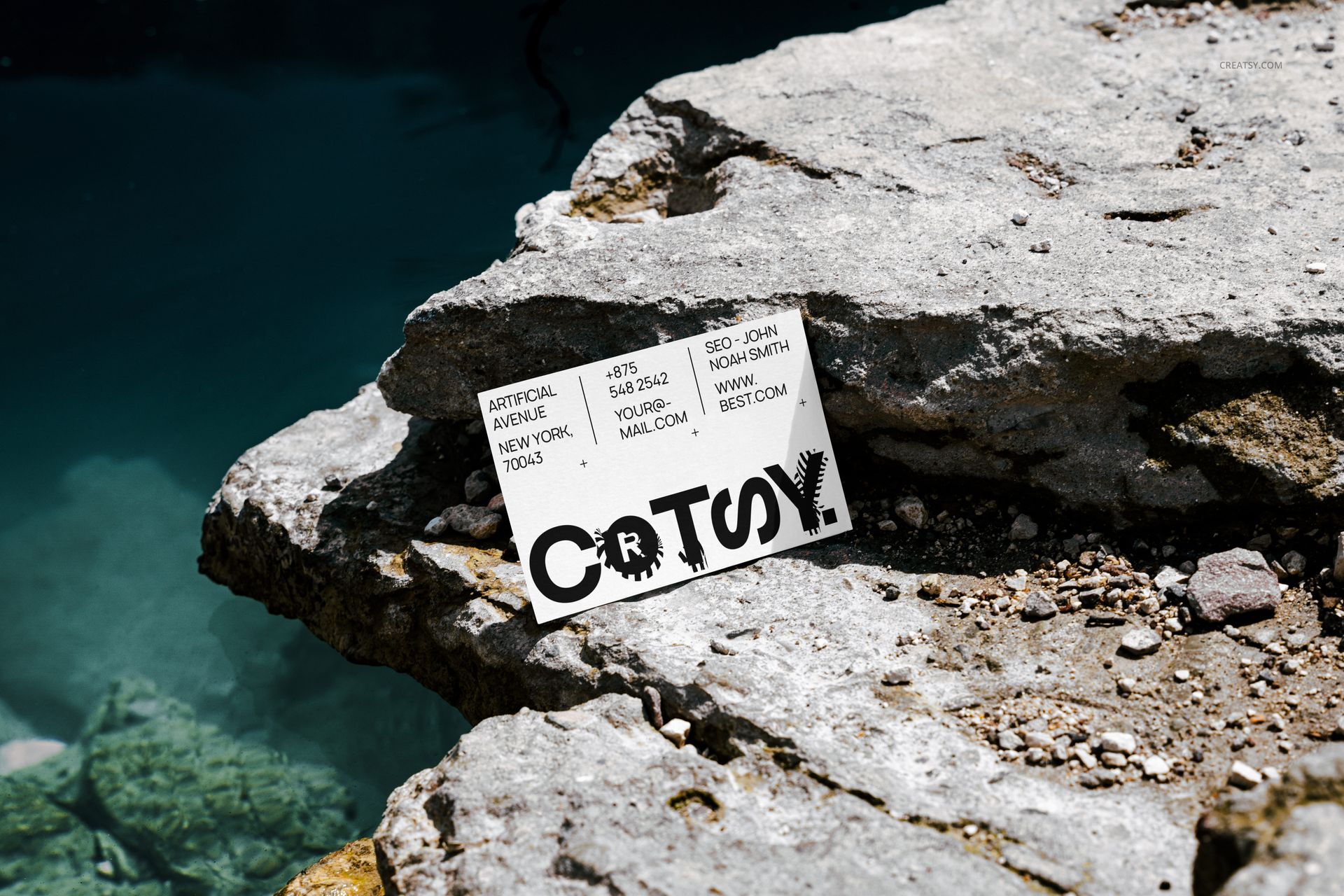Business card mockup placed on weathered stone near clear blue water, viewed from an angled perspective outdoors.