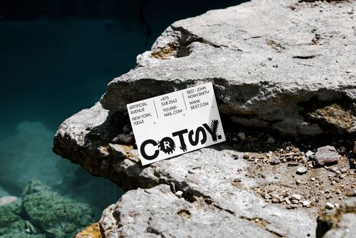Business card mockup placed on weathered stone near clear blue water, viewed from an angled perspective outdoors.