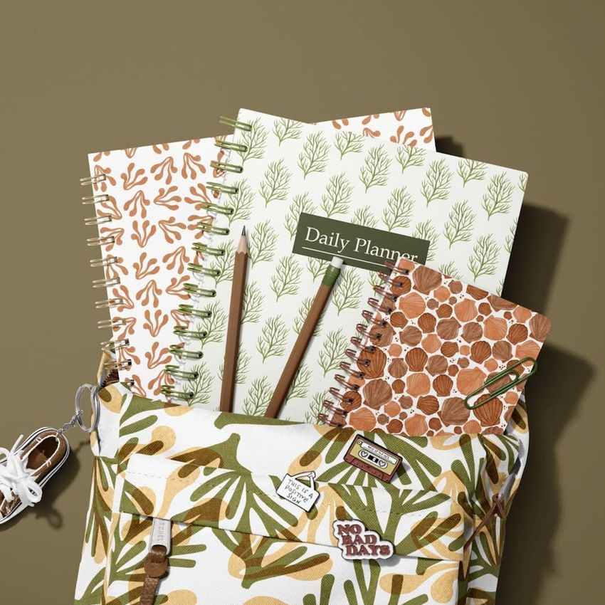 School backpack mockup with spiral notebooks featuring seaweed and coral patterns, pencils, and pins on an olive background.