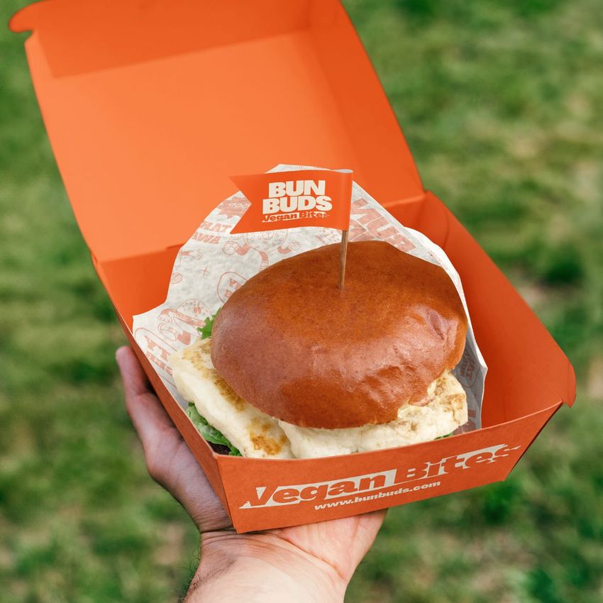 Vegan burger mockup in an open orange box with printed wrapper, held outdoors with blurred grass behind.