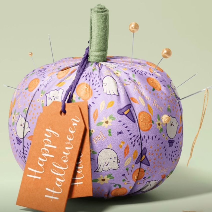 Fabric pumpkin mockup with a purple patterned surface featuring Halloween motifs, pins, and an orange gift tag on a plain background.