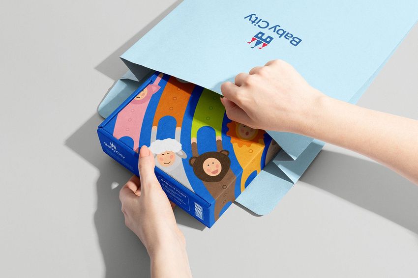 Hands placing a colorful illustrated box with cartoon animals into a light blue kraft mailer on a neutral background.