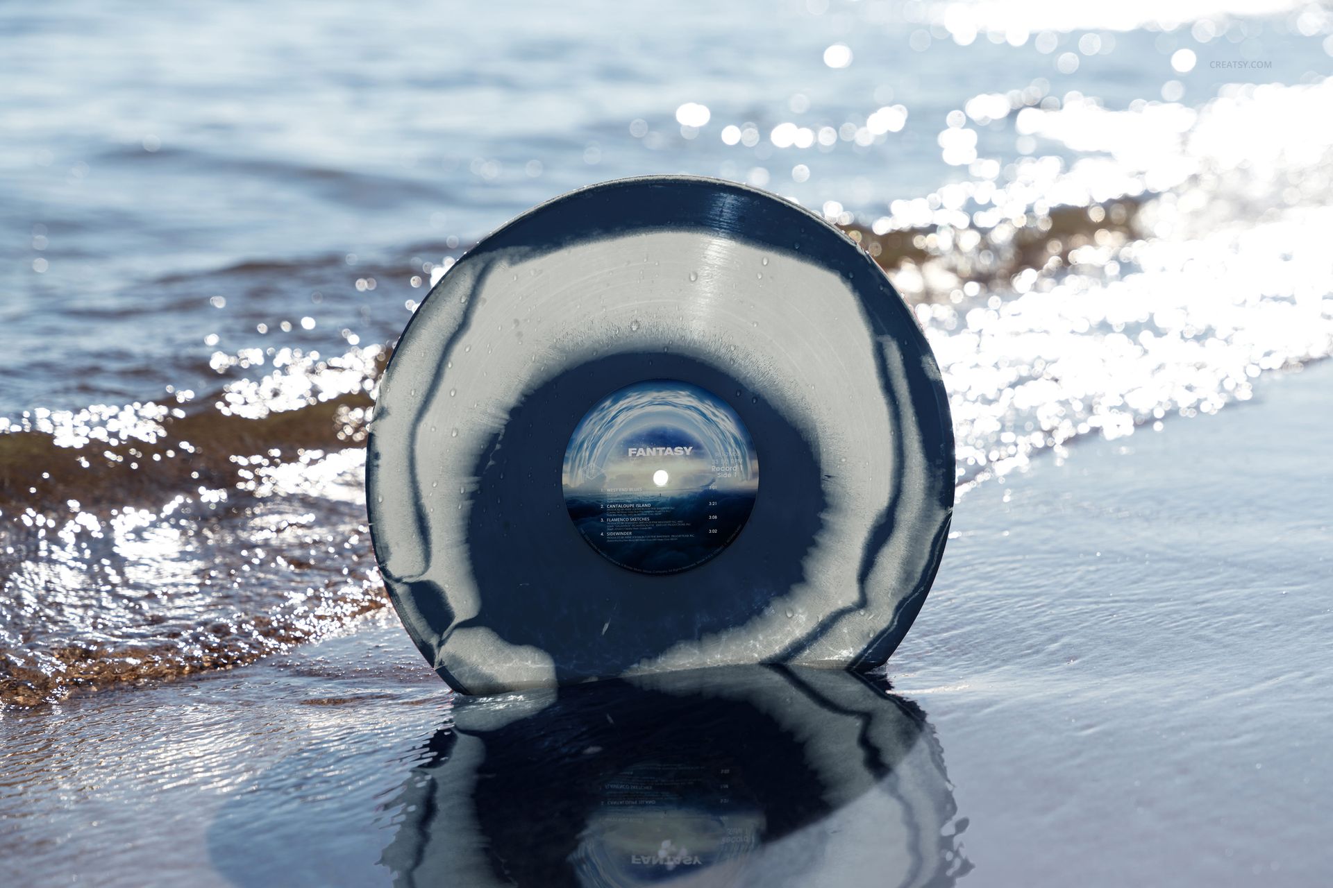 Mockup of a marbled vinyl record in shades of gray and black positioned on sandy beach by gentle waves.