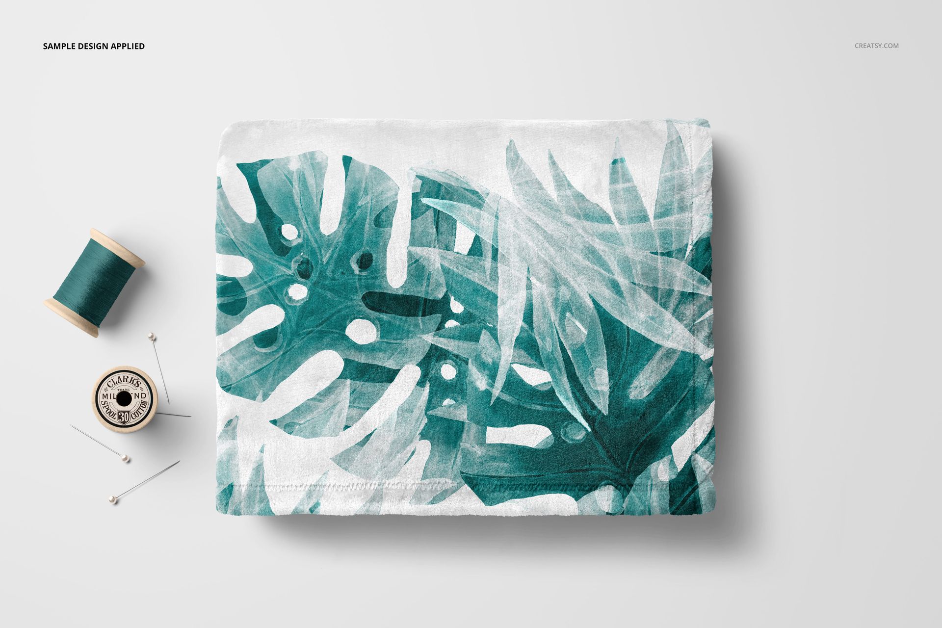 Fleece blanket mockup featuring large green leaves and white accents, folded neatly with sewing props nearby.