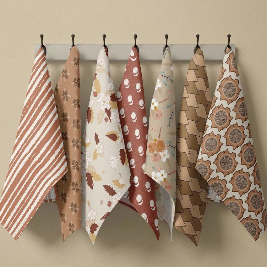 Hanging tea towels mockup showing six towels with various brown and beige patterns, displayed on black hooks against a neutral wall.