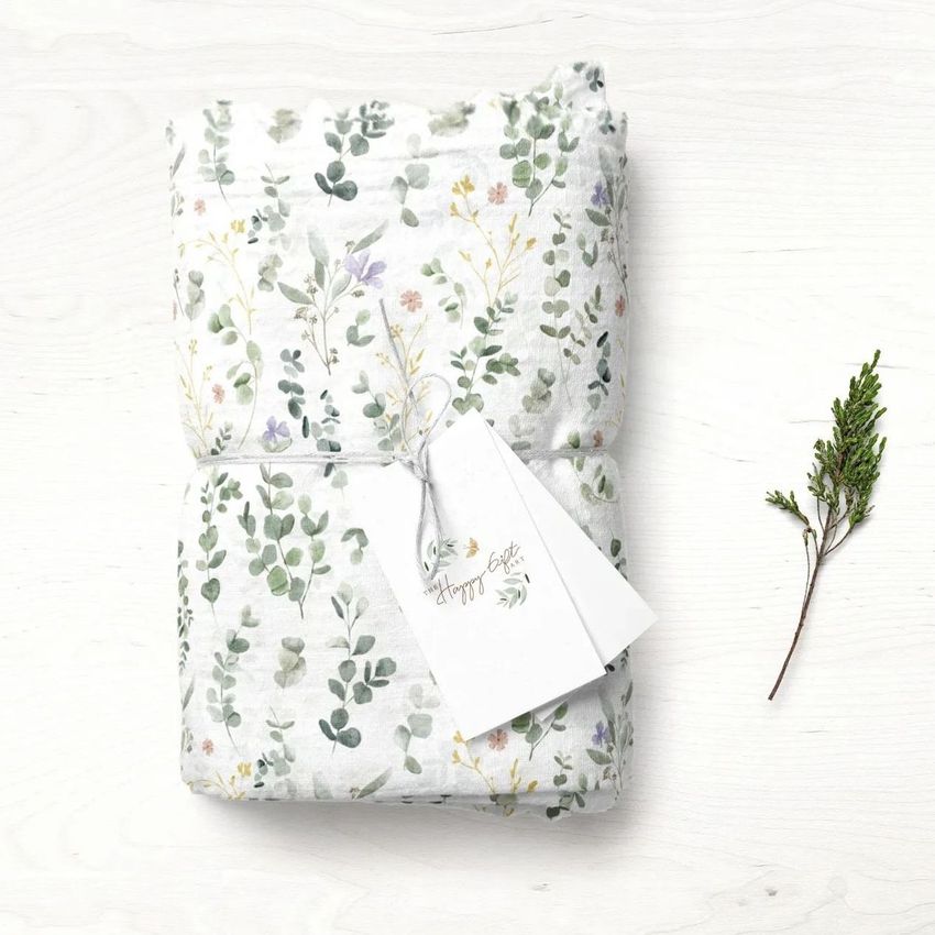 Muslin swaddle blanket mockup featuring a folded white blanket with a delicate eucalyptus and floral pattern, shown on a light wooden surface with a sprig and a blank gift tag attached.