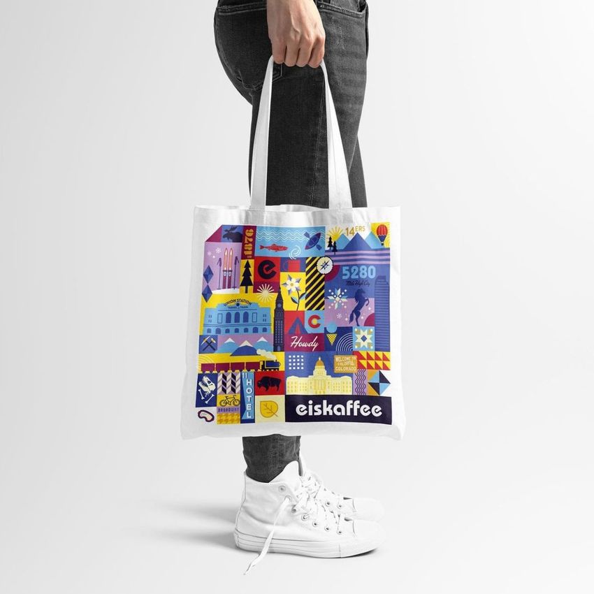 Tote bag mockup featuring a hand holding a white bag with a colorful geometric cityscape design and bold text at the bottom.
