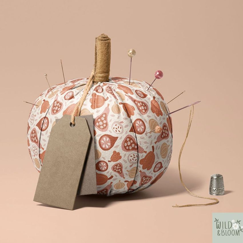 Fabric pumpkin mockup with a stitched stem, decorated with autumn leaf and pumpkin patterns, pins, a blank tag, and a thimble on a beige background.