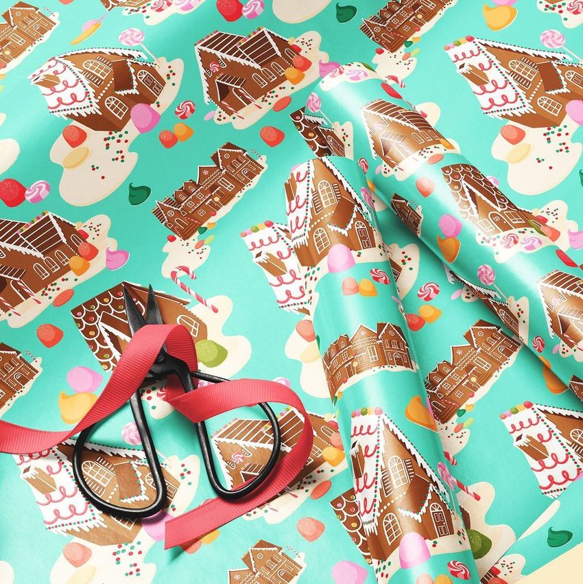 Gift wrapping paper mockup featuring a close-up of gingerbread house illustrations on a teal background with scissors and ribbon.