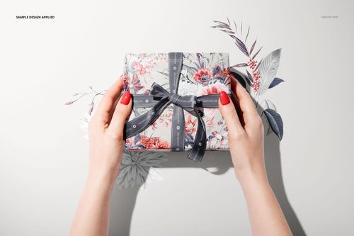 Top view of a gift box mockup wrapped in floral paper and tied with a dark ribbon, held by two hands.