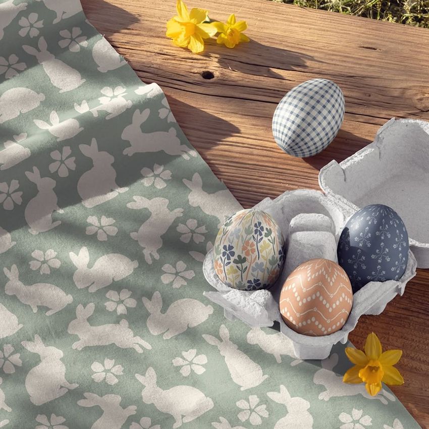 Cotton fabric mockup with white bunny and flower patterns on a green background, next to decorated Easter eggs in a carton.
