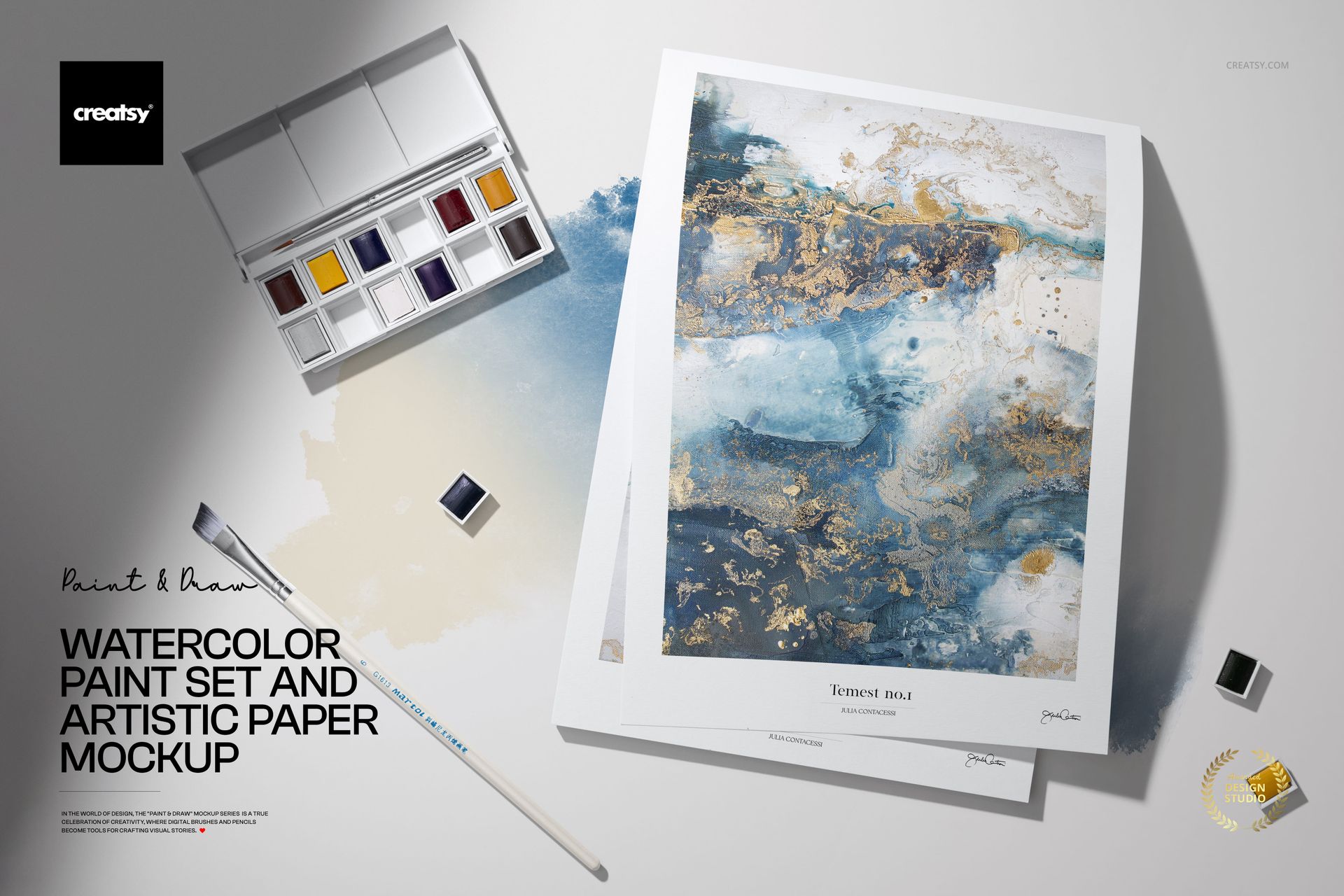 Watercolor paint set with open palette, paintbrush, and artistic paper featuring abstract blue and gold artwork, top view.