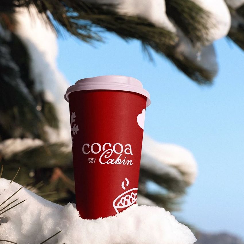 Red paper cup mockup with white winter-themed graphics and text, placed on snow with pine branches and blue sky in background.