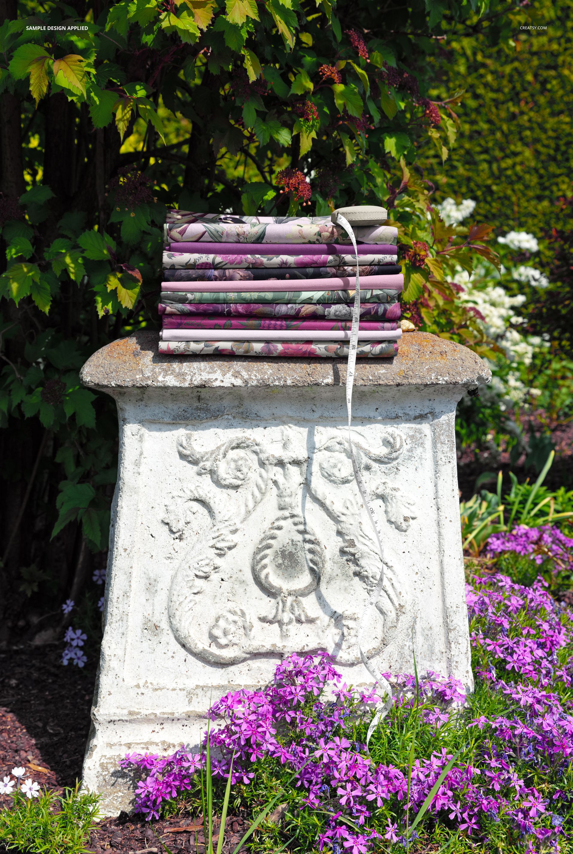 PSD mockup showing folded fabrics arranged on a carved stone pedestal outdoors, with vibrant greenery and flowers.