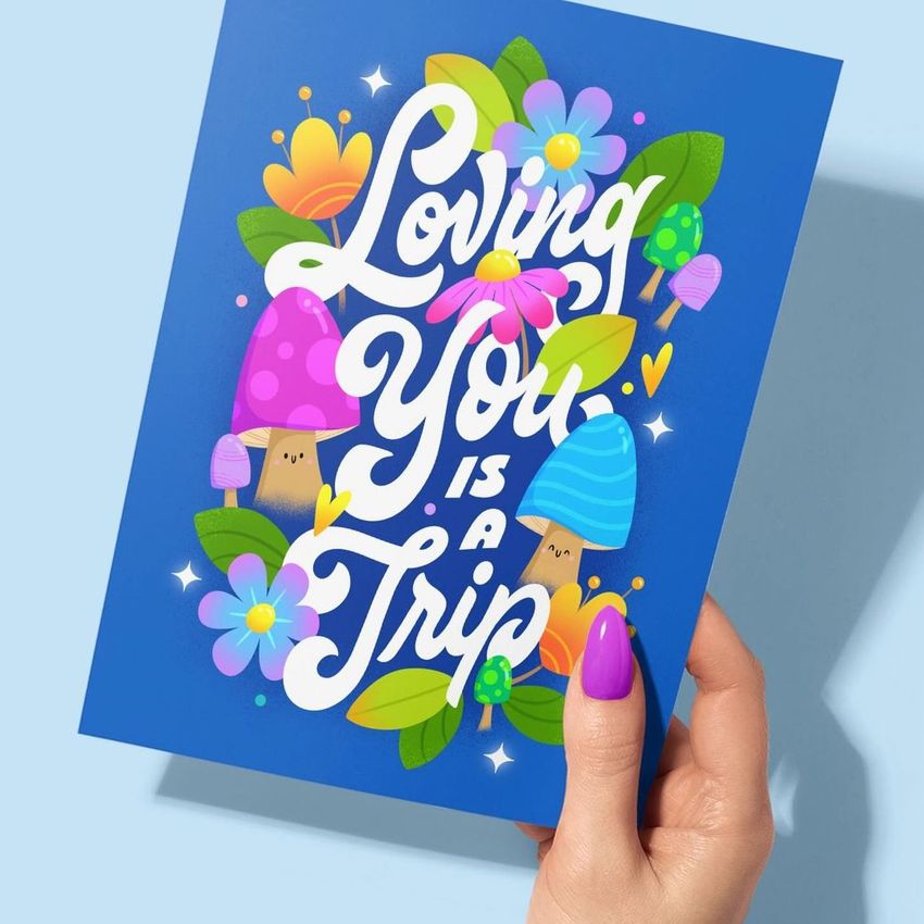 Greeting card mockup featuring a hand with purple nails holding a blue card decorated with colorful mushrooms, flowers, and bold text.