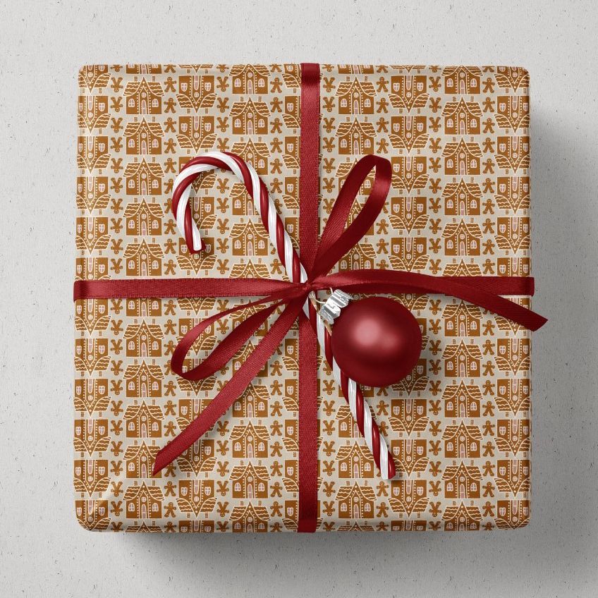 Christmas gift mockup featuring a rectangular box wrapped in patterned paper with gingerbread house motifs, tied with a red ribbon, and decorated with a candy cane and red ornament on a light surface.