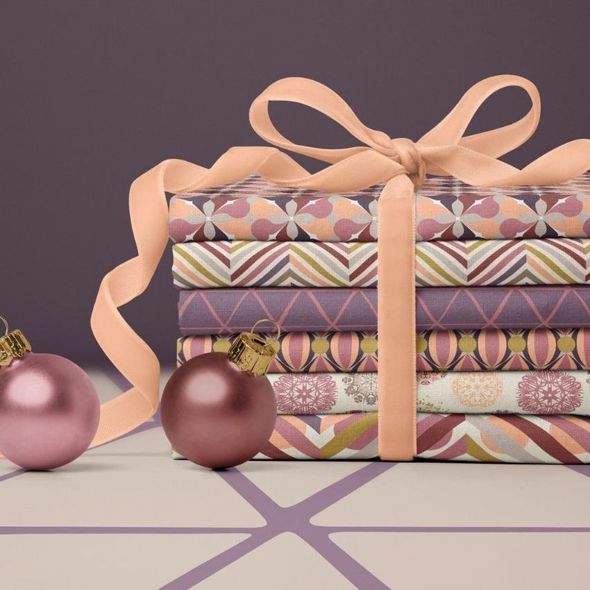 Fabric stack mockup featuring folded patterned fabrics tied with a peach ribbon, two pink Christmas baubles, and a purple background.