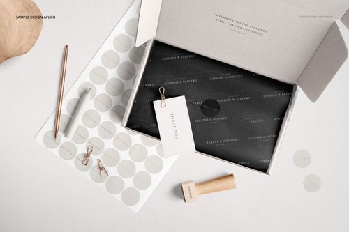 Open mailing box mockup with black tissue paper, thank you card, pen, stamp, and round sticker sheets on a light surface.