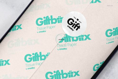 Gift box mockup showing white wrapping tissue paper with teal graphics and a round sticker, seen from a top angle.
