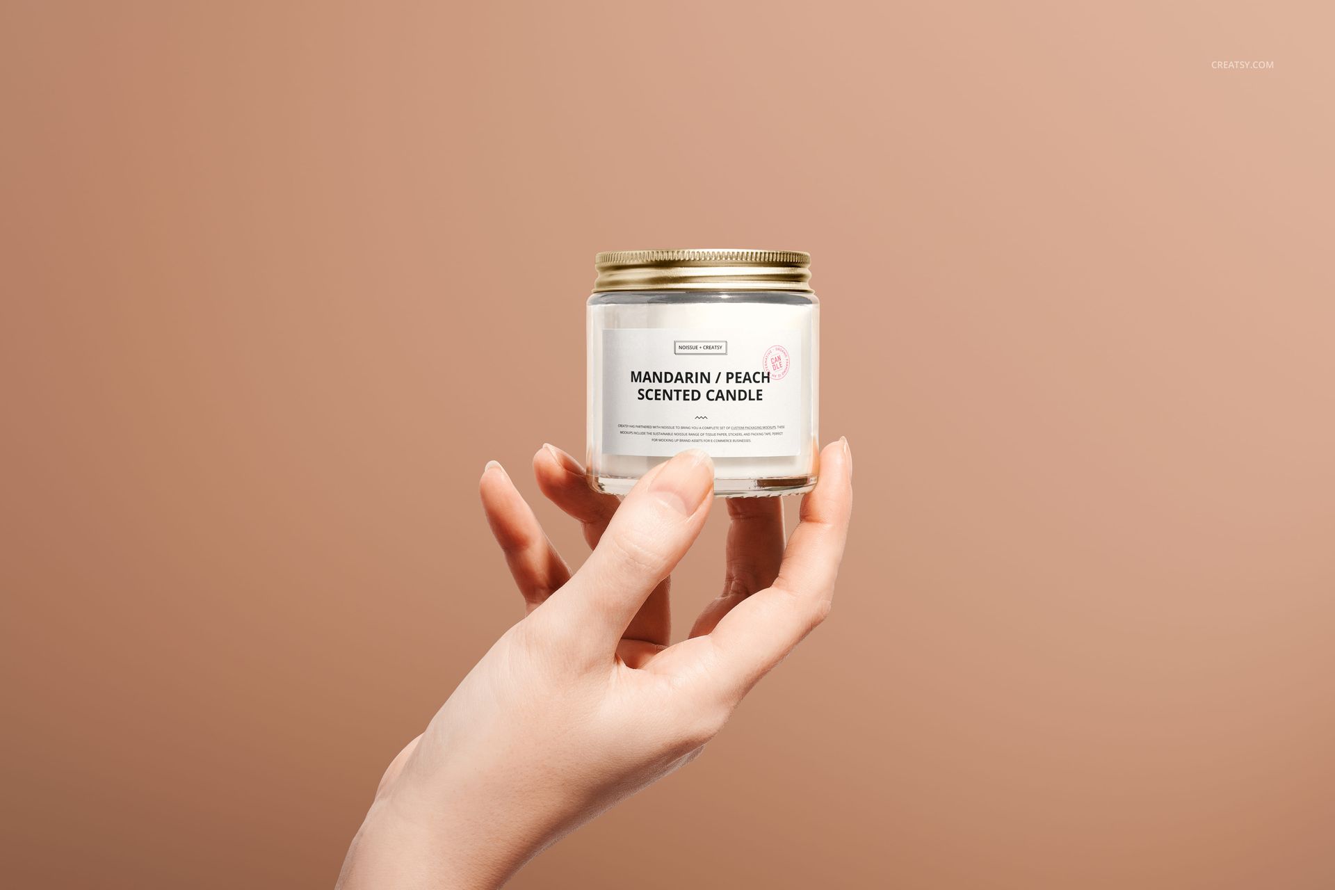 Mockup of a jar candle featuring a white label and gold lid, displayed in a hand with a plain beige background.