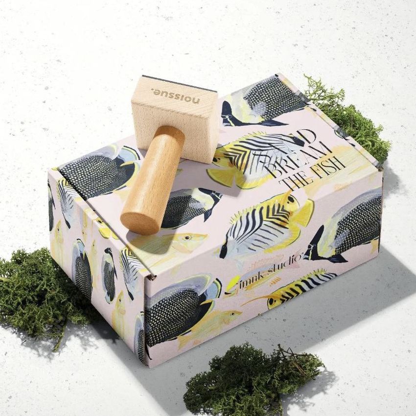 Wooden stamp mockup resting on a pastel box decorated with colorful fish illustrations, surrounded by green moss on a light surface.