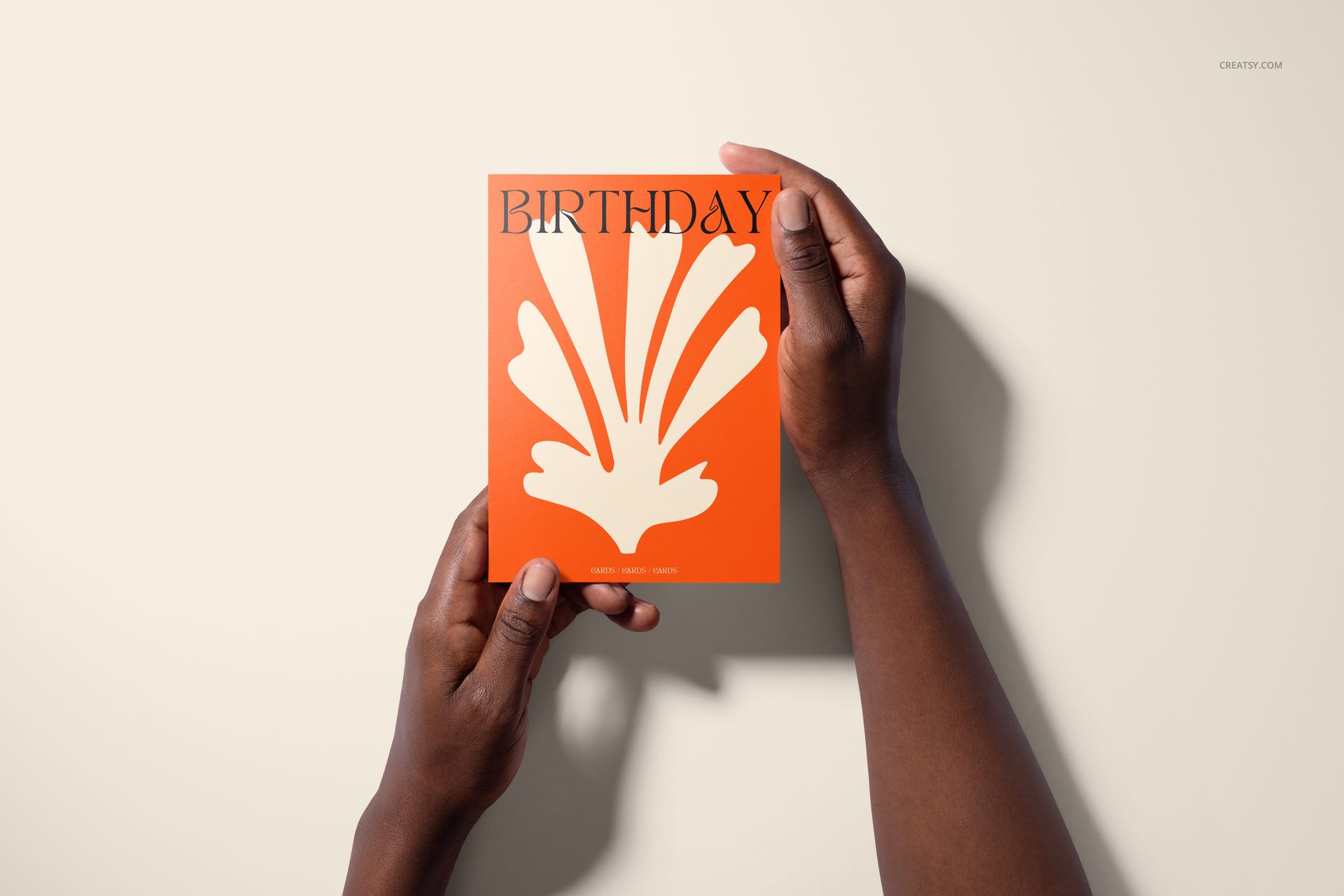 Top-down view of two hands displaying a birthday greeting card with orange cover and white decorative motif, mockup scene.