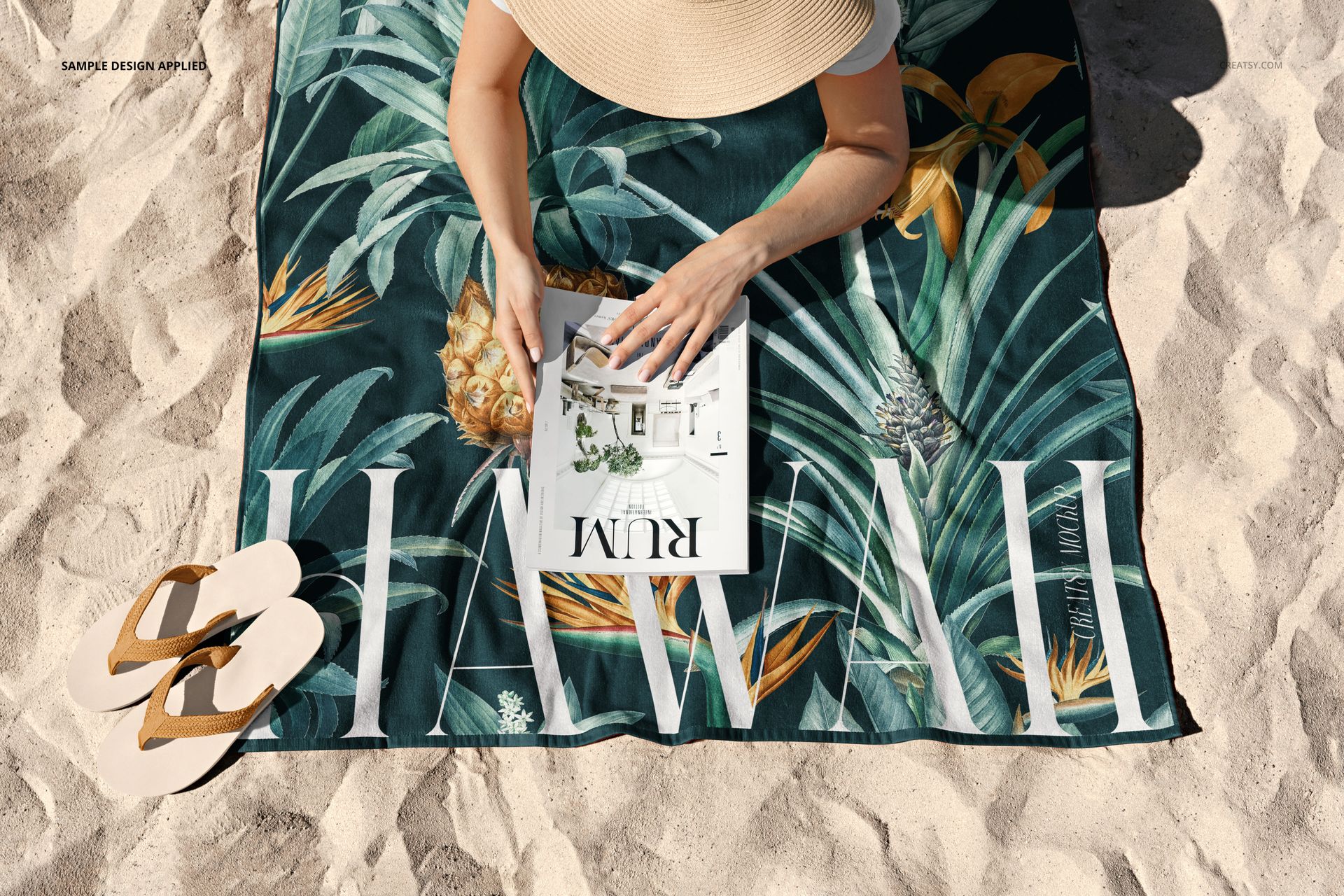 Beach towel mockup featuring green foliage and pineapple design, person in sun hat reading on sandy beach.