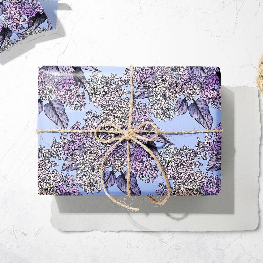 Gift wrapping paper mockup featuring a rectangular box wrapped in floral patterned paper with purple and lilac flowers, tied with twine on a light surface.
