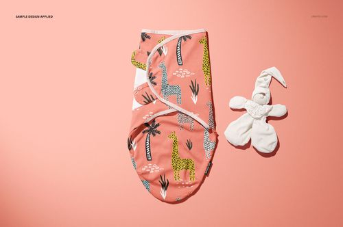 Top view of a baby swaddle wrap mockup featuring a pink fabric with giraffe and tree prints, next to a white plush toy.