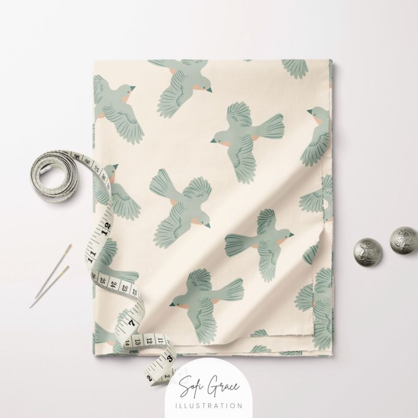 Folded cotton fabric mockup featuring a bird pattern in muted green on a cream background, shown with a measuring tape and sewing pins.