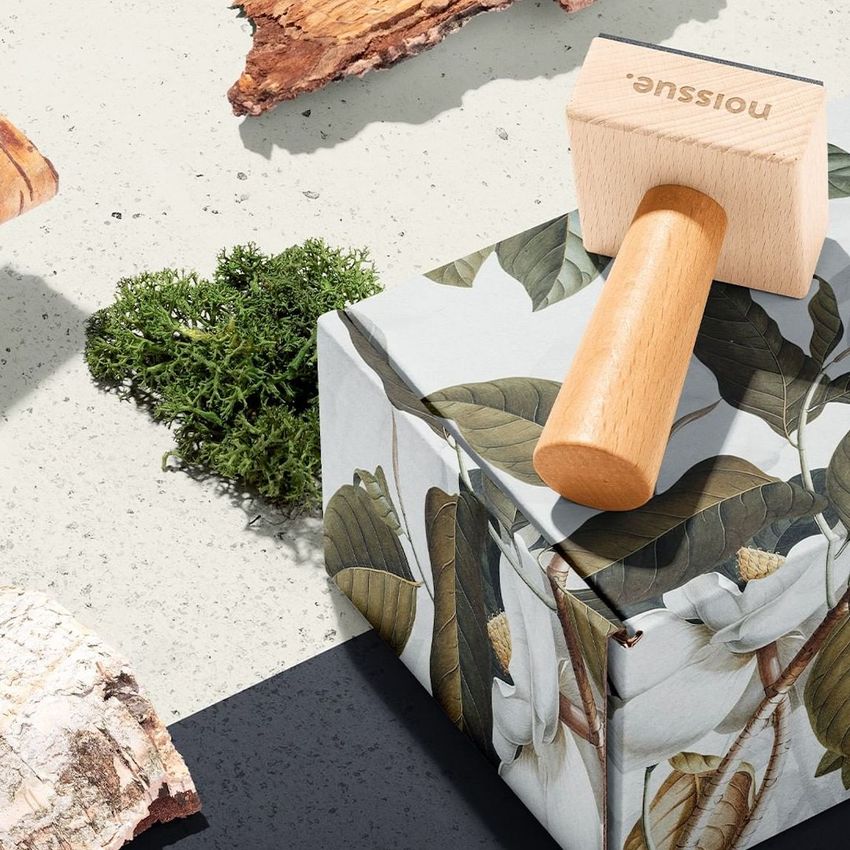 Wooden stamp mockup resting on a floral patterned box, surrounded by pieces of bark and green moss on a neutral surface.