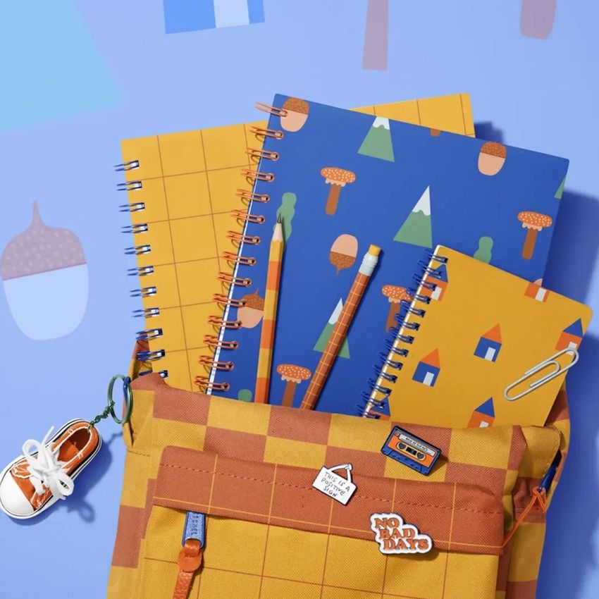 School backpack mockup with spiral notebooks in orange and blue patterns, pencils, and a sneaker keychain on a blue surface.