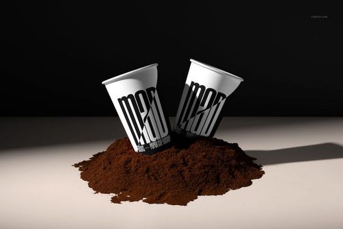 PSD mockup showing two paper cups with black lettering, half-buried in ground coffee, illuminated by strong directional light.