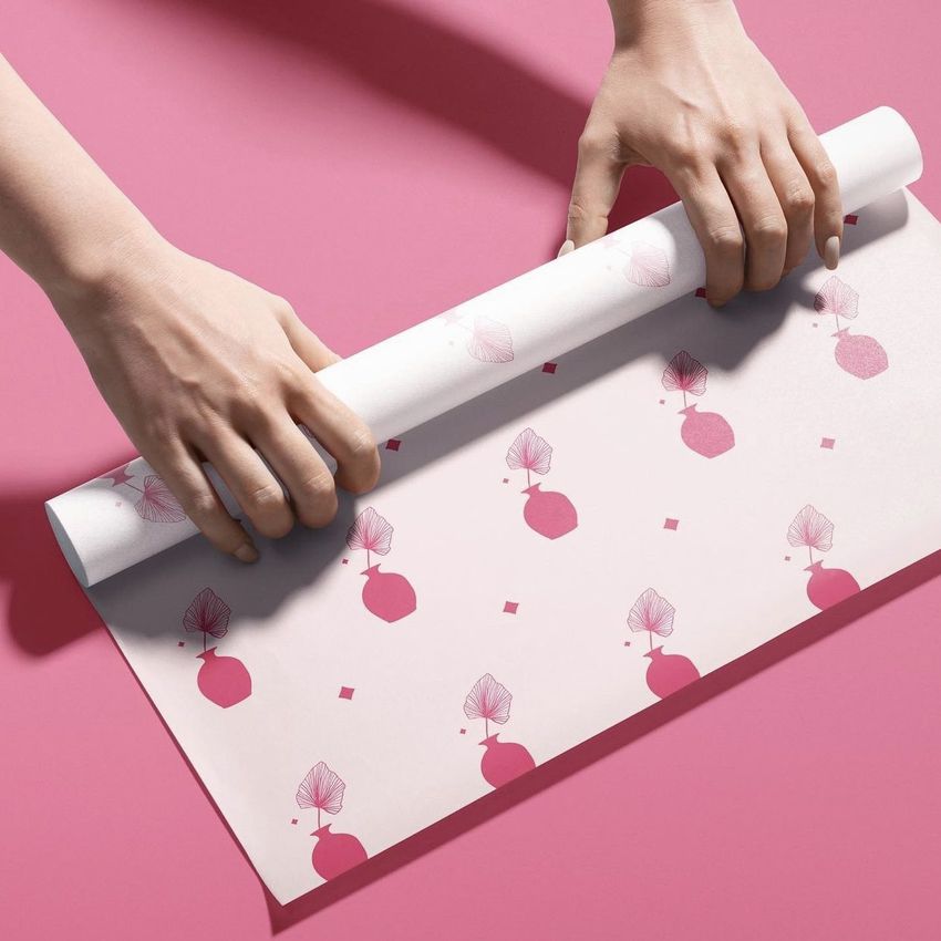 Hands unrolling a sheet of food safe paper mockup with pink geometric and leaf patterns on a solid pink background.