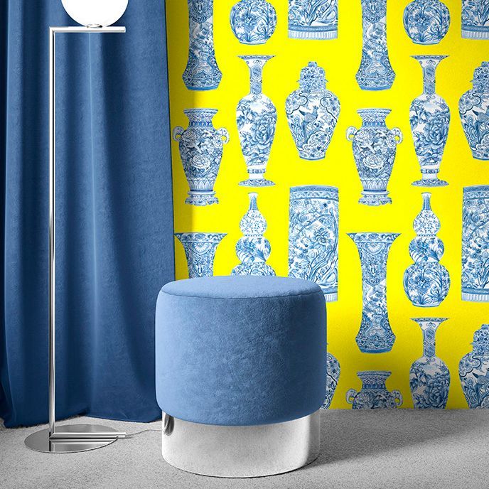 Interior scene mockup showing a blue velvet stool on a gray floor, next to a chrome lamp and blue curtain, with a yellow wall featuring blue and white vase illustrations.