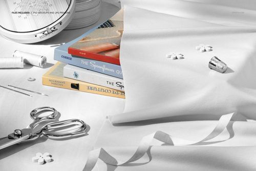 Flat lay of a sewing area featuring white jersey fabric mockup, metallic scissors, thimble, and stacked books.