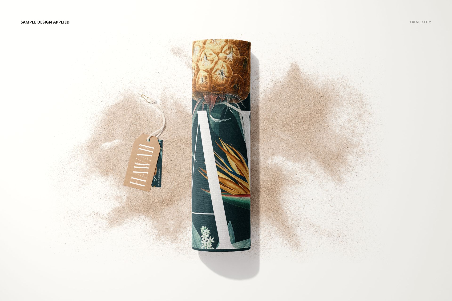 Rolled beach towel mockup with tropical leaf and pineapple pattern, placed on scattered sand with a brown tag attached.