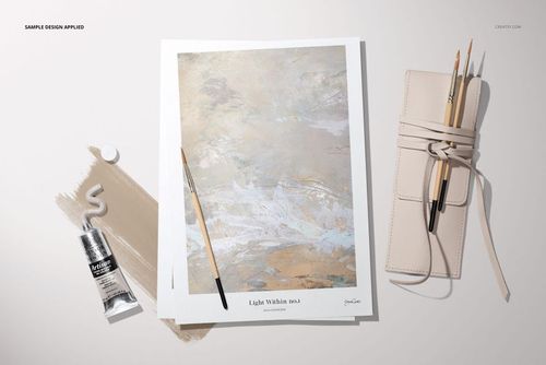 Top view of art paper with abstract painting, paintbrush, beige paint tube, and rolled-up brush holder on a light surface.