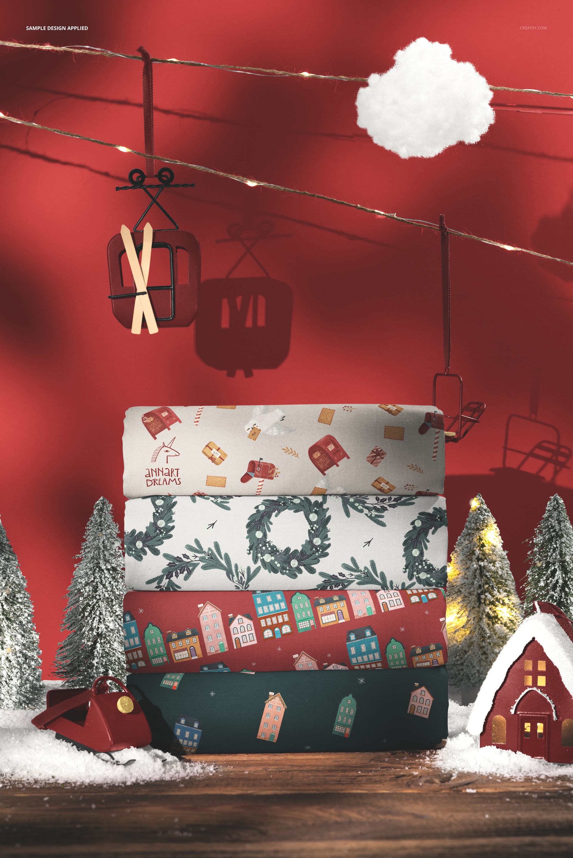 Christmas scene mockup showing stacked cotton fabrics with holiday designs, surrounded by miniature trees and decorations.