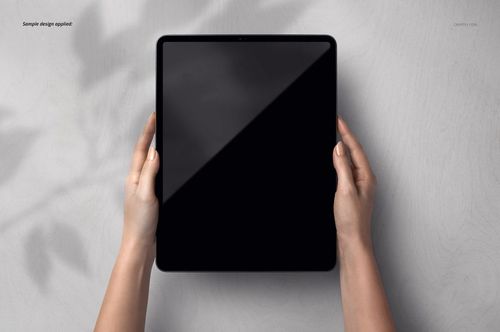 Hands holding a black screen iPad Pro tablet viewed from above on a light gray wooden surface in a mockup scene.