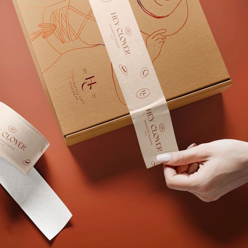 Kraft mailing box mockup on a warm brown surface, decorated with minimalist line art and branded paper tape being applied by hand.