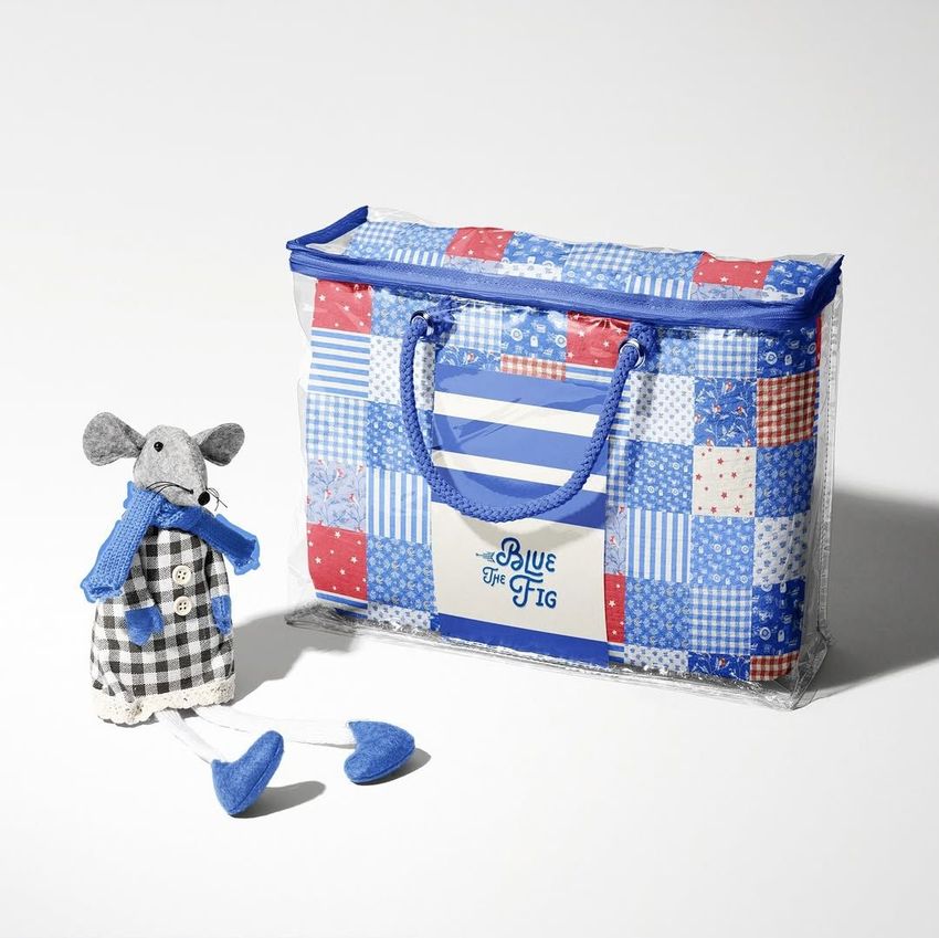 Newborn swaddle wrap mockup featuring a folded quilt in a clear zippered bag with blue handles and a plush mouse toy beside it.