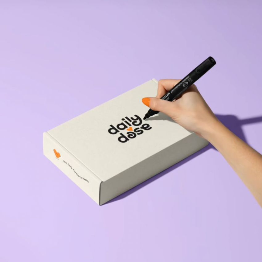 Mailing box mockup on a light purple surface with a hand writing on the lid, featuring bold black text and an orange accent.