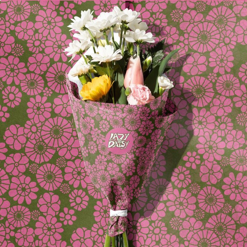 Flowers packaging mockup showing a bouquet wrapped in pink floral-patterned paper with a visible text label, set against a matching patterned background.