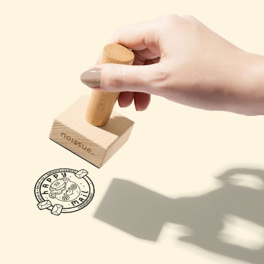 Hand holding a wooden stamp mockup pressing a circular stamp design with text and small icons onto a light beige surface.